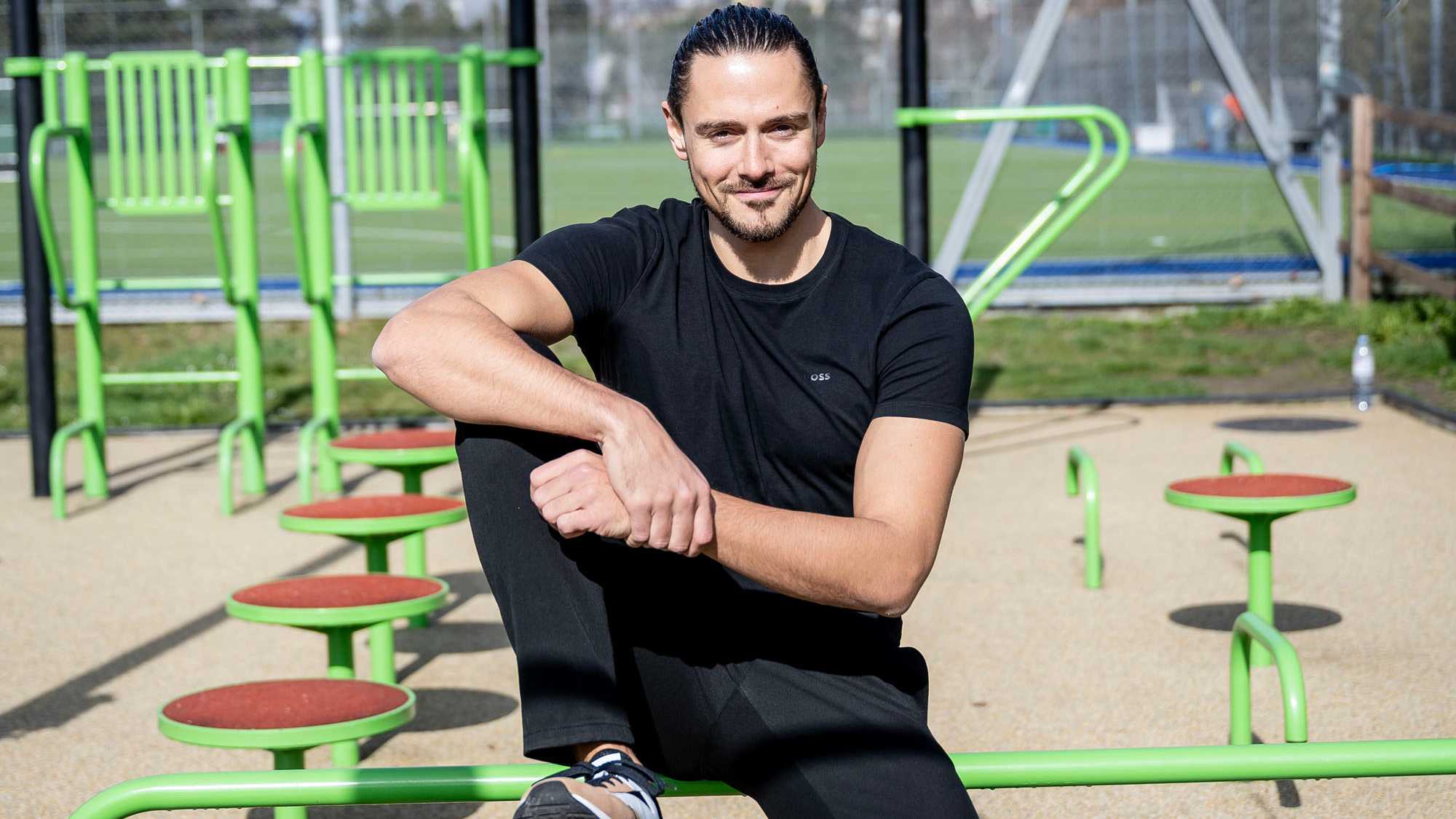 Joel Roos, casually dressed in black, sitting outdoors on some bright green sports equipment 